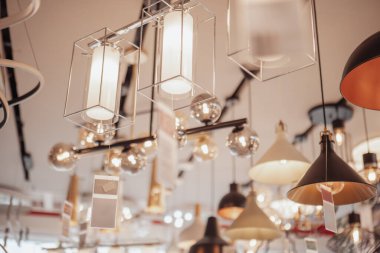 Kolye ışıkları, geometrik çerçeveler ve küresel ampuller de dahil olmak üzere çeşitli tasarımlar, şekiller ve stiller sergilenen bir mağazada çeşitli modern aydınlatma armatürleri sergilenmektedir..