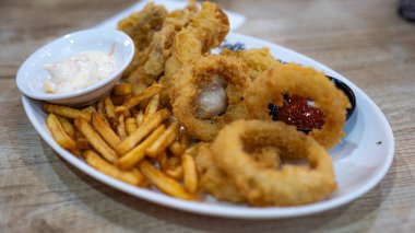 Kızarmış soğan halkaları, altın patates kızartması ve bir kase kremalı sosla dolu beyaz bir tabağın yakın çekimi. Lezzetli bir aperatif ya da meze için mükemmel, taze ve sıcak servis edilir..