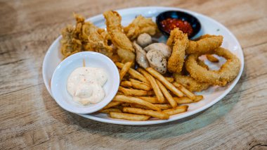 Kızarmış soğan halkaları, altın patates kızartması ve bir kase kremalı sosla dolu beyaz bir tabağın yakın çekimi. Lezzetli bir aperatif ya da meze için mükemmel, taze ve sıcak servis edilir..