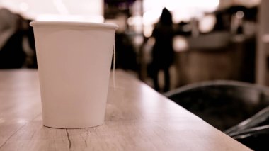 Kafe ortamında, arka planda bulanık bir şekilde duran ahşap bir masada çay poşeti ipiyle beyaz bir kağıt bardak. Resmi ve metni atayabileceğiniz düz kağıt bardaklar.