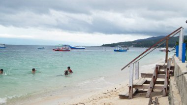 Donggala, Central Sulawesi 'de kumlu bir sahil. Sakin sulara demir atmış birkaç tekne ve kıyıya yakın yüzen birkaç kişi var. Tahta bir merdiven sahile iniyor. Gökyüzü bulutlarla kaplı..