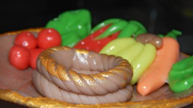 Resim, domatesler, yapraklar, muzlar, bir havuç ve altın aksanlı bükülmüş kahverengi bir halka da dahil olmak üzere renkli kil gıda modellerini gösteriyor..