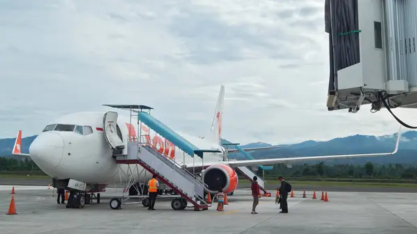 Pistteki Boeing 737-800 Lion Air. Seyyar merdivenden inen yolcularla birlikte. Havaalanı personeli, güvenlik konileri ve bir jet köprüsü mevcut, bulutlu bir gökyüzünün altında dağ gibi bir arka planı var..