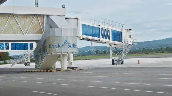 Görüntü Mutiara Havaalanı 'ndaki bir havaalanı jet köprüsünü gösteriyor. Jet köprüsü terminali uçağa bağlıyor ve yolcu alımını kolaylaştırıyor. Arka planda bir pist ve uzak tepeler var..