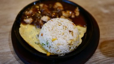 Görüntü, bir tabak yemek, bir porsiyon kızarmış pirinç, bir omlet ve koyu soslu bir et gösteriyor. Pirinç bir kubbeye dönüşür ve tabak siyah bir tabakta servis edilir..