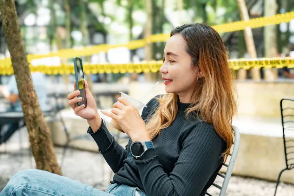 Siyah elbiseler ve kot pantolon giyen güzel bir kadın elinde soğuk bir fincan kahveyle açık bir kafede oturuyor ve cep telefonuyla iletişim kuruyor..