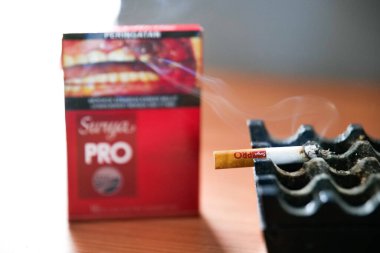 The lit cigarette butt is placed in the ashtray, and behind it is the cigarette packet. Gudang Garam Surya brand.