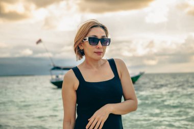 Omuz hizasında sarı saçlı ve güneş gözlüklü çok güzel bir kadın öğleden sonra sahilde eğleniyordu. Gökyüzü altın sarısı ve deniz berrak. Arkasında bir balıkçı teknesi vardı..