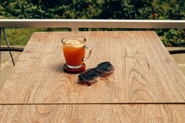 Üzerinde portakal dilimi olan bir bardak zerdeçal içeceği, ahşap bir masada servis edilir. Yanında siyah bir gözlük var.. 