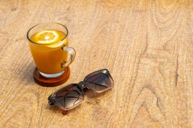 Üzerinde portakal dilimi olan bir bardak zerdeçal içeceği, ahşap bir masada servis edilir. Yanında siyah bir gözlük var.. 