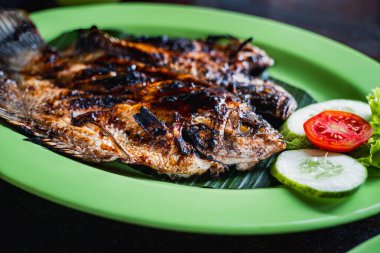 Yeşil bir tabakta ızgara sazan, salatalık ve domates dilimleri ve altında marul ile servis edilir..