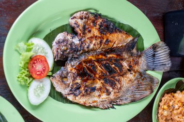 Yeşil bir tabakta ızgara sazan, salatalık ve domates dilimleri ve altında marul ile servis edilir..