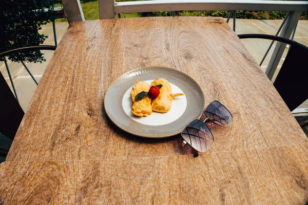 Açık bir kafede ahşap bir masa. Üzerinde, kızarmış yiyecekler seramik tabakta servis ediliyor. Yanında güneş gözlüğü var..