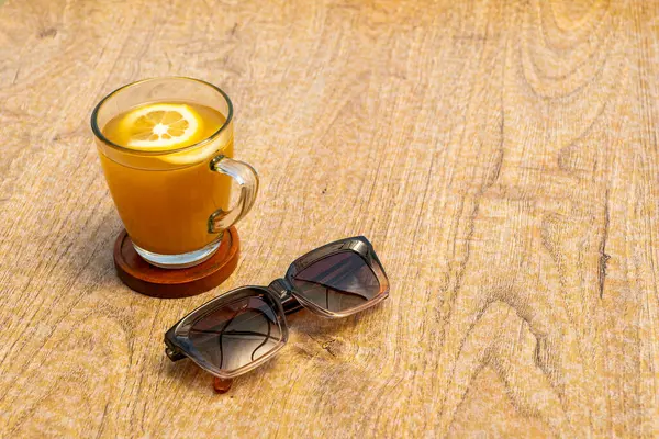 Üzerinde portakal dilimi olan bir bardak zerdeçal içeceği, ahşap bir masada servis edilir. Yanında siyah bir gözlük var.. 