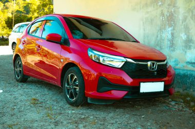 Evin yanına park edilmiş kırmızı Honda Brio marka küçük bir araba. Beyaz bir duvar ve pencerenin yanında..