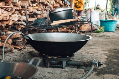 Açık bir alanda yemek pişirmek için yere konmuş bir gaz sobası. Üzerinde büyük bir tava ile düzgünce dizilmiş odun yığınının arka planı.