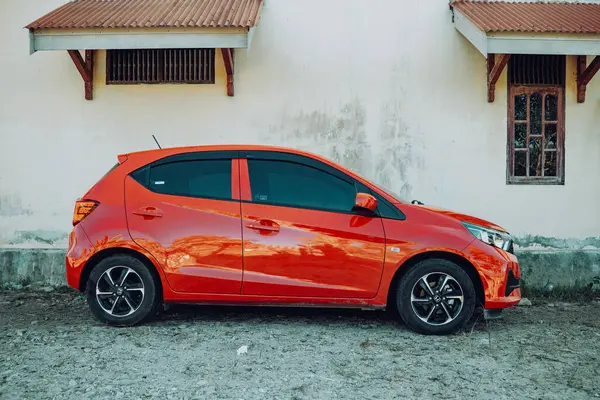 Evin yanına park edilmiş kırmızı Honda Brio marka küçük bir araba. Beyaz bir duvar ve pencerenin yanında..