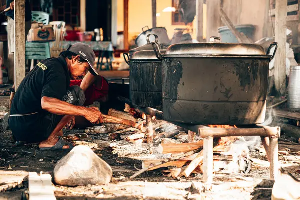 Bir adam büyük bir kazanda yemek pişirmek için odun yakıyor..
