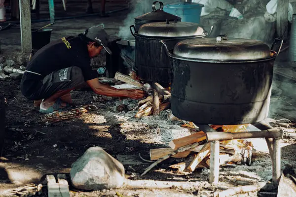 Bir adam büyük bir kazanda yemek pişirmek için odun yakıyor..