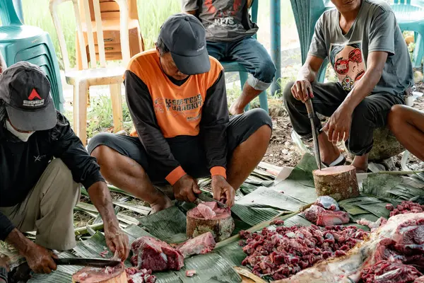 Bir grup adam yere çömelmiş, yere konmuş muz yapraklarıyla domuz eti kesiyor..