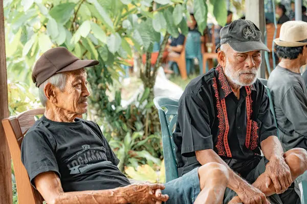 Bir grup adam oturuyor ve konuşuyor. Arkalarında pirinç tarlaları ve dağlar var. Ayrıca odun yığını..