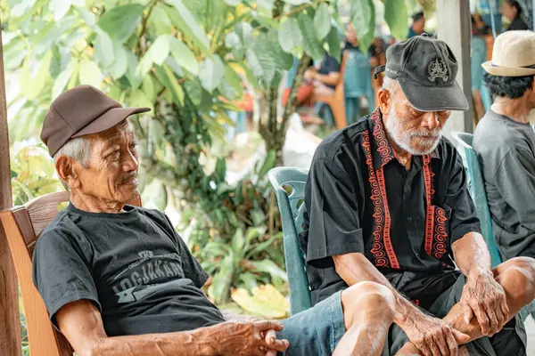 Bir grup adam oturuyor ve konuşuyor. Arkalarında pirinç tarlaları ve dağlar var. Ayrıca odun yığını..