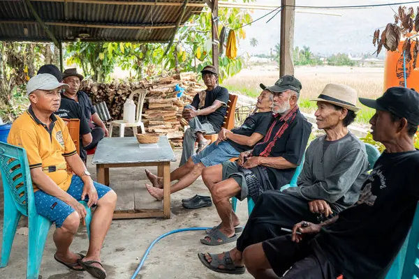 Bir grup adam oturuyor ve konuşuyor. Arkalarında pirinç tarlaları ve dağlar var. Ayrıca odun yığını..