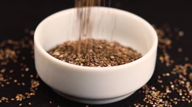 Chia tohumları siyah arka planda beyaz bir seramik kaseye düşüyor.