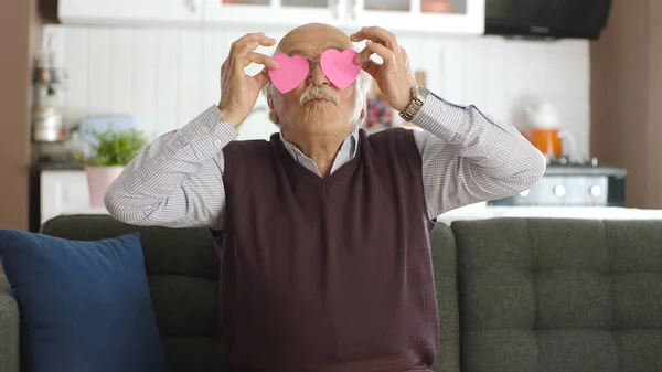 Happy old man playing with two paper hearts in his eyes. Old man sending kisses to his wife on sofa at home with message photo with red paper heart. Valentine's day, love in eyes, romantic concept.