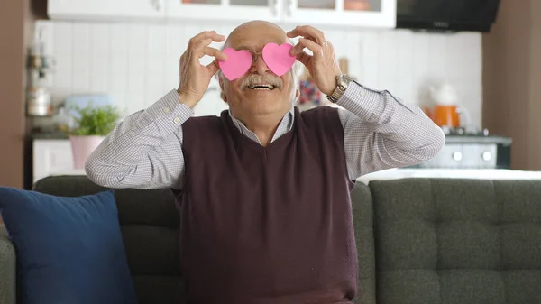 Happy old man playing with two paper hearts in his eyes. Old man sending message photo with red paper heart to his wife on sofa at home. Valentine\'s day, love in eyes, romantic concept.