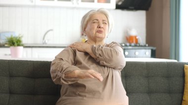 Yaşlı kadın boyun eklemlerinde ağrı hisseder. Kanepede oturan yaşlı kadının omzu ve sırt ağrısı var. Ağrıyan bölgelerine masaj yapan bir kadın. Eklem sorunları olan yaşlı bir kadının portresi.