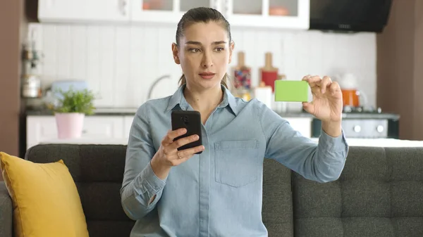 Woman holding smartphone and green box on her sofa, showing a product, smiling and presenting an imaginary object. Creative 3d artists can replace the green box with any product they want.