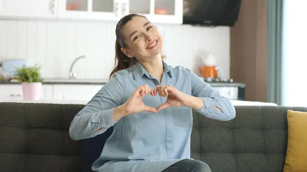 Young woman smiling and making heart sign at camera with hands at home. Healthy heart health and love concept.Valentine's day,mother's day,romantic concept. Heart health insurance, world heart day.
