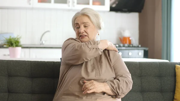 Elderly woman feels pain in neck joints. Elderly woman sitting on sofa has shoulder and back pain. Woman massaging her aching areas. Portrait of an elderly woman with joint problems.