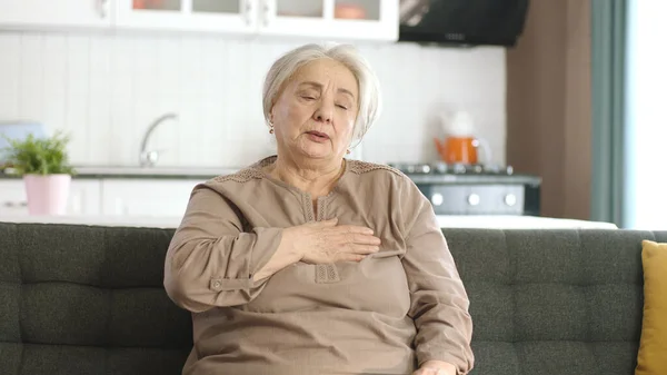 Elderly woman feels pain in her chest. Elderly woman sitting in her armchair at home is having a heart attack. Portrait of old woman with heart palpitations. Acute chest pain, heart attack.
