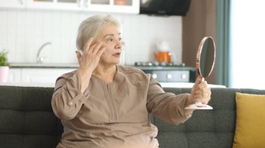 Son sınıf öğrencisi kadın aynaya bakarken cildini ve saçlarını kontrol ediyor. Mutlu gülümseyen, kendini şımartan, sağlıklı cilt bakımı, yaşlanan güzellik, cilt bakımı kozmetik ürünler konsepti.