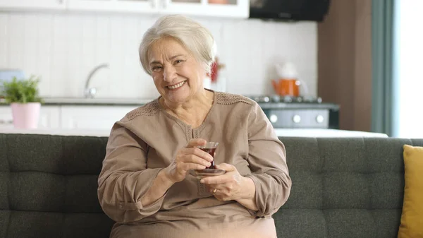 Happy old woman watching an interesting movie on TV while drinking tea. The old woman is sitting on the sofa, watching a mysterious TV movie alone.Entertainment. Old humans. Feelings.