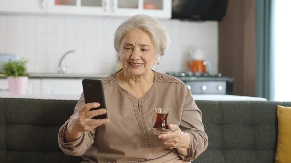 快乐的老奶奶拿着手机 一边喝茶一边和心爱的人视频通话 一位老年妇女正在学习使用现代技术设备 老年人使用的技术 — 图库照片