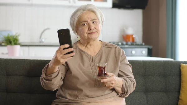 快乐的老祖母拿着手机 听到她一边喝茶一边和心爱的人打视频电话时 非常惊讶 一位老年妇女正在学习使用现代技术设备 — 图库照片