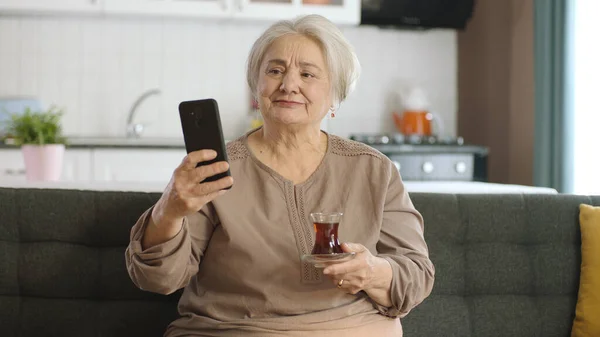 快乐的老祖母拿着手机 听到她一边喝茶一边和心爱的人打视频电话时 非常惊讶 一位老年妇女正在学习使用现代技术设备 — 图库照片
