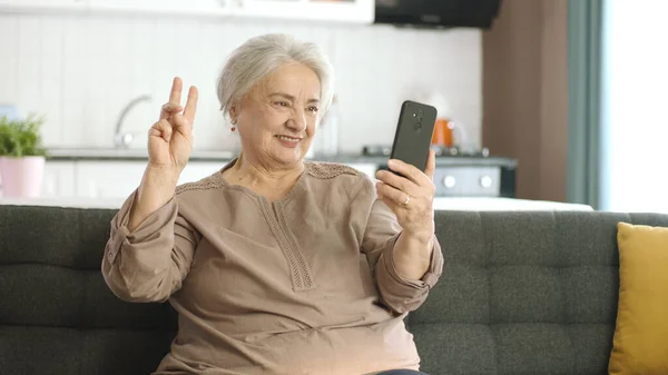 老妇人笑着 和亲戚们一起打视频电话 看着相机挥手 坐在舒适的沙发上 与老年人一起使用技术的概念 带着胜利标志的自拍 — 图库照片