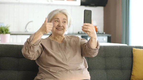 Senior woman smiling, enjoying video call with relatives, looking at camera and waving, sitting on comfortable sofa. Technology use concept with the elderly. Taking selfie with thumbs up sign.