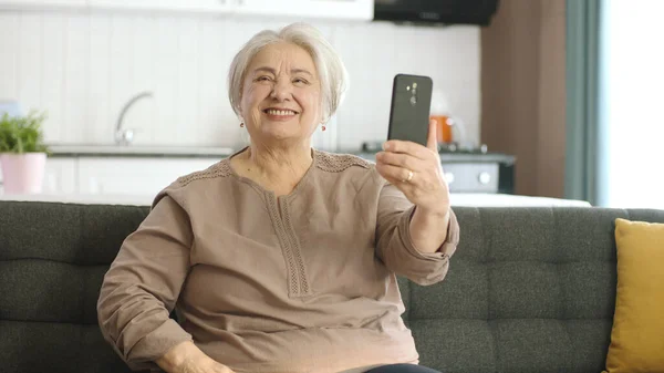 老妇人笑着 和亲戚们一起打视频电话 看着相机挥手 坐在舒适的沙发上 老年人的技术使用概念 — 图库照片