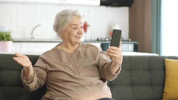 老妇人笑着 和亲戚们一起打视频电话 看着相机挥手 坐在舒适的沙发上 老年人的技术使用概念 — 图库照片
