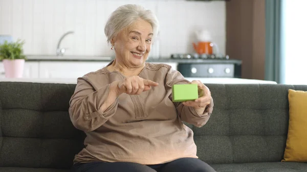 这位老妇人在扶手椅上拿着绿色的东西 展示着一种产品 微笑着 展示着一种愉快的 想象的物体 有创意的3D艺术家可以用他们想要的任何产品来代替绿色的盒子 — 图库照片