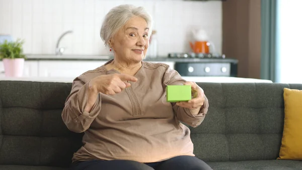 Yaşlı Kadın Koltuğunda Yeşil Bir Şey Tutuyor Ürünü Gösteriyor Gülümsüyor — Stok fotoğraf
