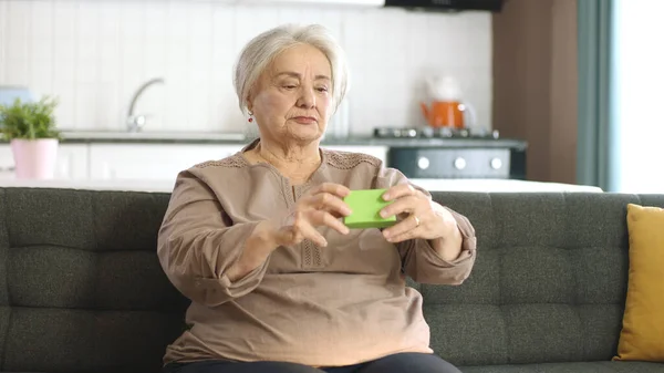 这位老妇人在扶手椅上拿着绿色的东西 展示着一种产品 微笑着 展示着一种愉快的 想象的物体 有创意的3D艺术家可以用他们想要的任何产品来代替绿色的盒子 — 图库照片