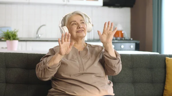 一位老妇人在沙发上一边听音乐 一边用耳机听音乐 有趣的老妇人带着耳机 唱歌跳舞 享受退休生活的成熟而快乐的女人 — 图库照片
