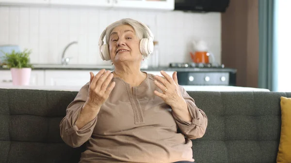 一位老妇人在沙发上一边听音乐 一边用耳机听音乐 有趣的老妇人带着耳机 唱歌跳舞 享受退休生活的成熟而快乐的女人 — 图库照片