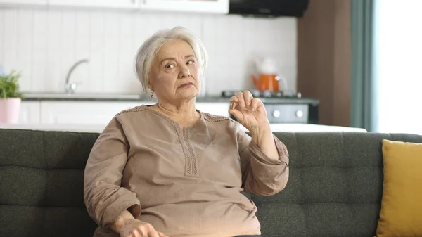 An elderly white-haired woman points to a pill on the living room sofa. She points out that the medicine in her hand is perfect. Happy old woman showing health care, disease treatment, vitamins.